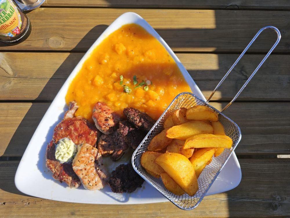 Sunlit plate on a wooden table with grilled mixed meats (including salmon and sausage), a creamy orange pumpkin mash topped with a few green onion slices, and a small metal basket of roasted potato wedges.