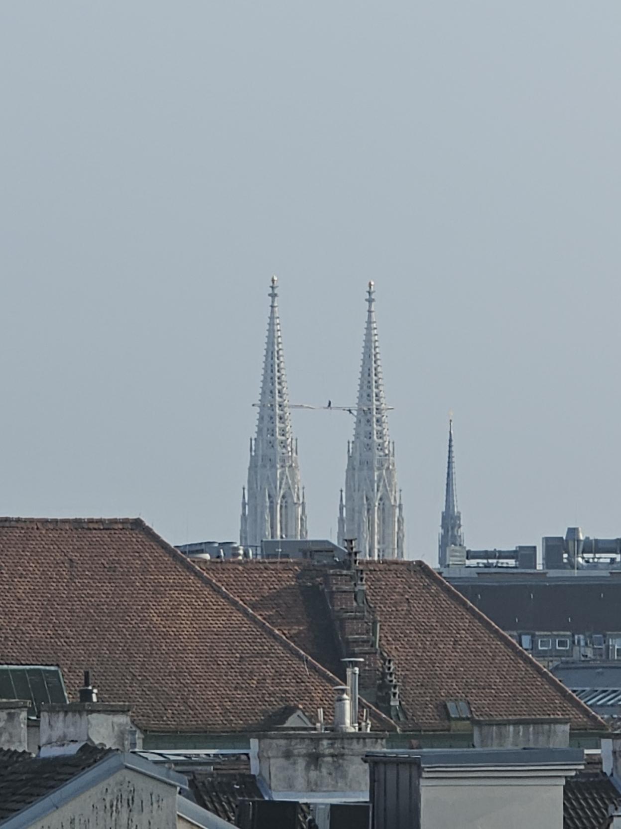 Zwei Arbeiter stehen hoch oben zwischen den Türmen der Wiener Votivkirche und arbeiten an einem beleuchteten Symbol in Form einer liegenden Acht vor dem hellen Himmel.