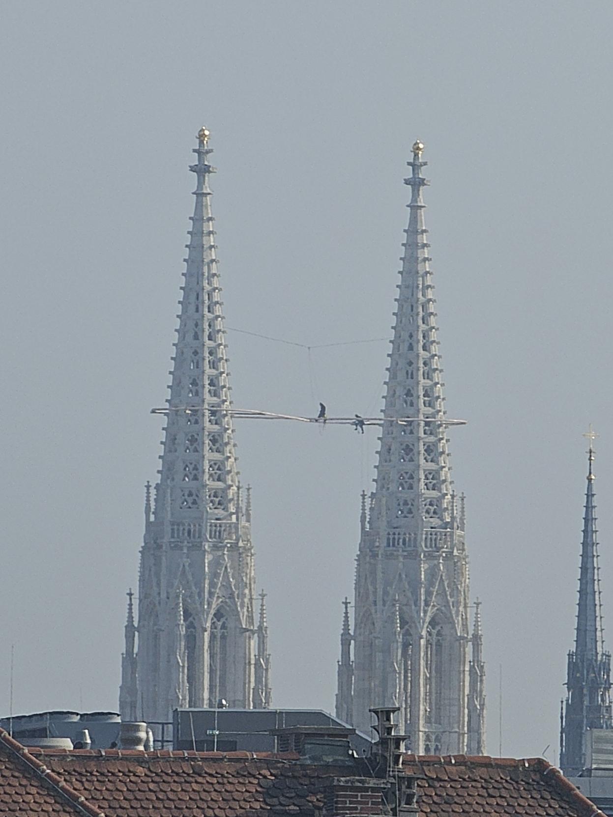 Zwei Arbeiter stehen hoch oben zwischen den Türmen der Wiener Votivkirche und arbeiten an einem beleuchteten Symbol in Form einer liegenden Acht vor dem hellen Himmel.