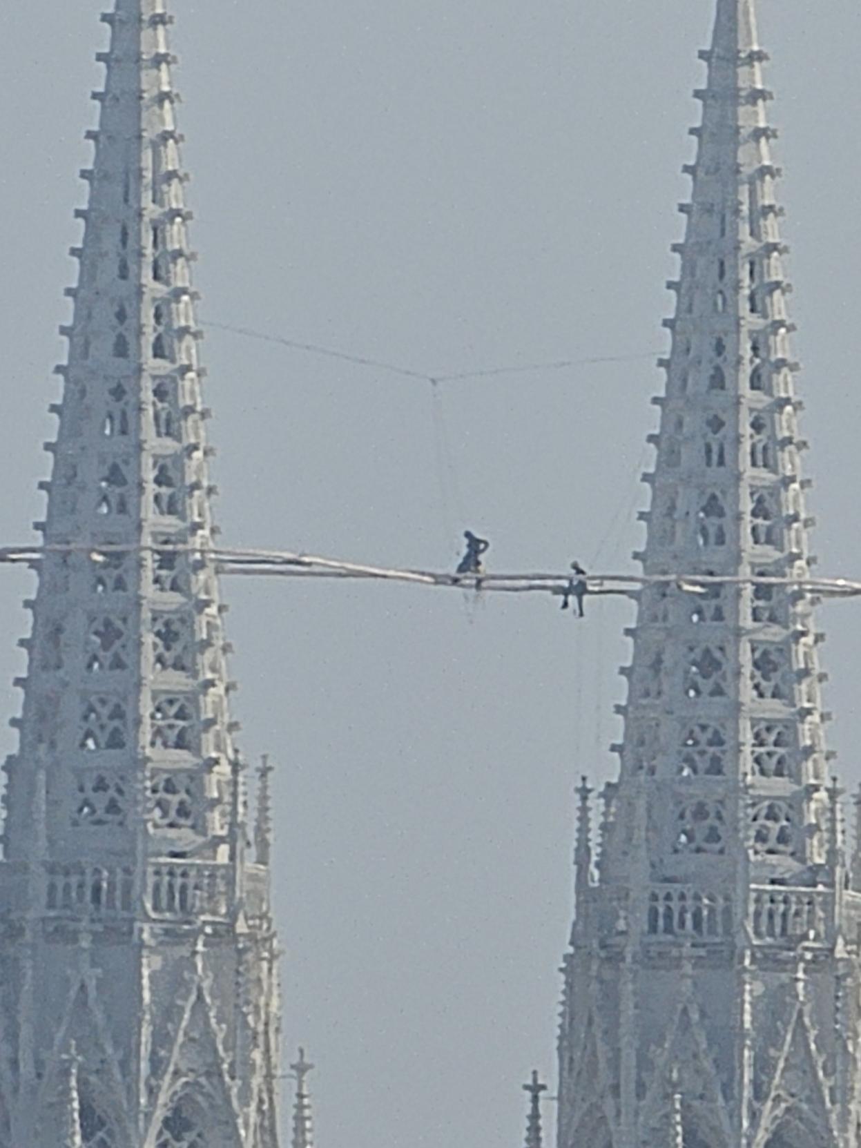 Zwei Arbeiter stehen hoch oben zwischen den Türmen der Wiener Votivkirche und arbeiten an einem beleuchteten Symbol in Form einer liegenden Acht vor dem hellen Himmel.